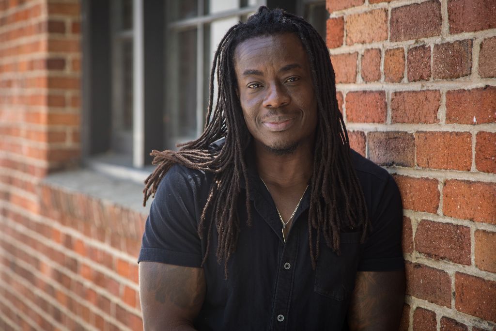 Skyler Cooper leaning against a brick wall, wearing a black short-sleeve button-up shirt.
