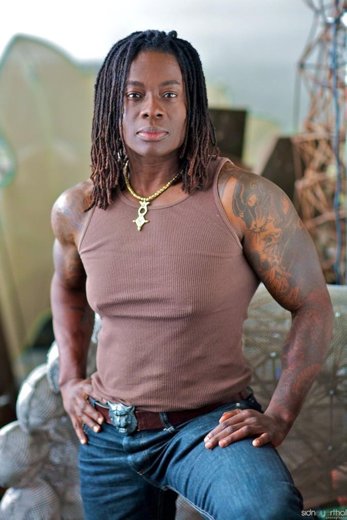 Skyler Cooper, standing confidently in a brown tank top with visible tattoos and a gold chain.